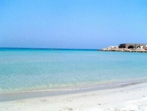 A beach with clear turquoise water and a sandy shore. There is a structure with arches in the background. - Residence Blue Sky (Melendugno)