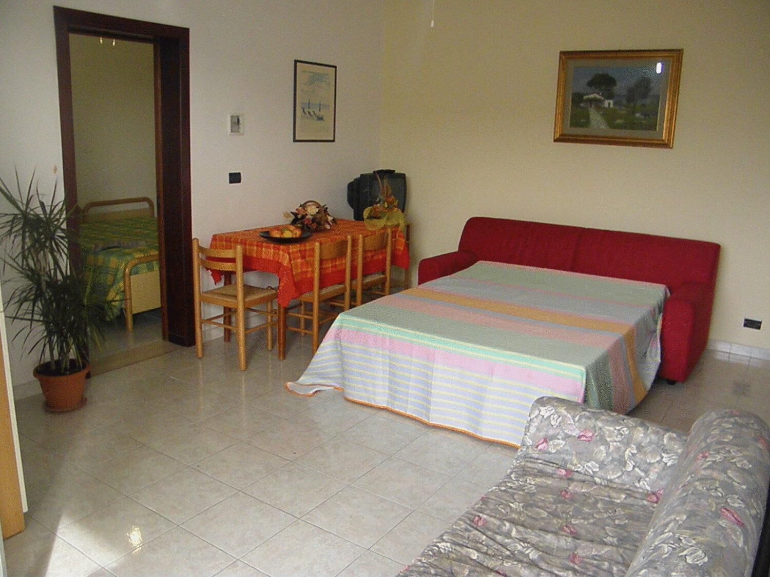 A room with a bed, a small table with chairs, a potted plant, and a painting on the wall.