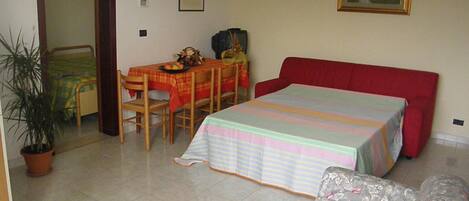 A room with a bed, a small table with chairs, a potted plant, and a painting on the wall.