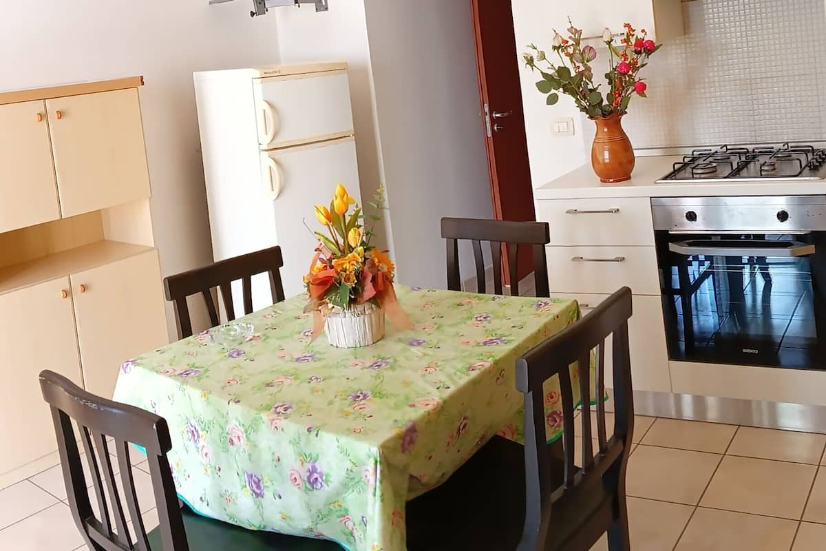 A dining area with a table covered in a floral tablecloth, surrounded by wooden chairs. A vase with flowers is placed on the table.