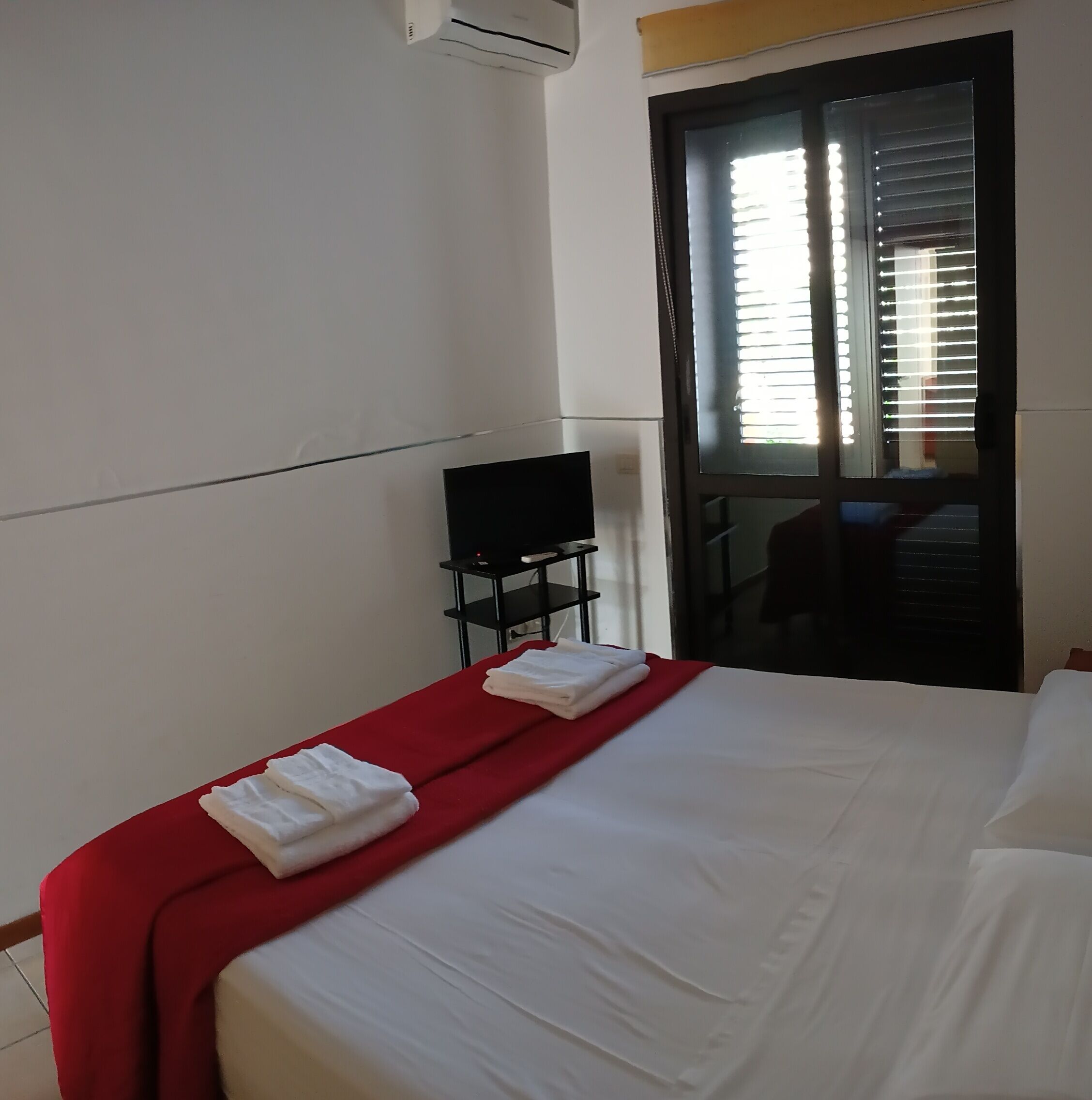 A hotel room with a bed, a TV on a small table, and a window with blinds.