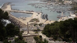 Uma marina costeira com barcos atracados, uma praia arenosa e um muro de pedra histórico.