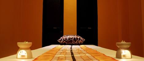 Habitación de hotel con una cama en el centro, dos velas encendidas, un cojín decorativo y vistas a una ventana oscura.