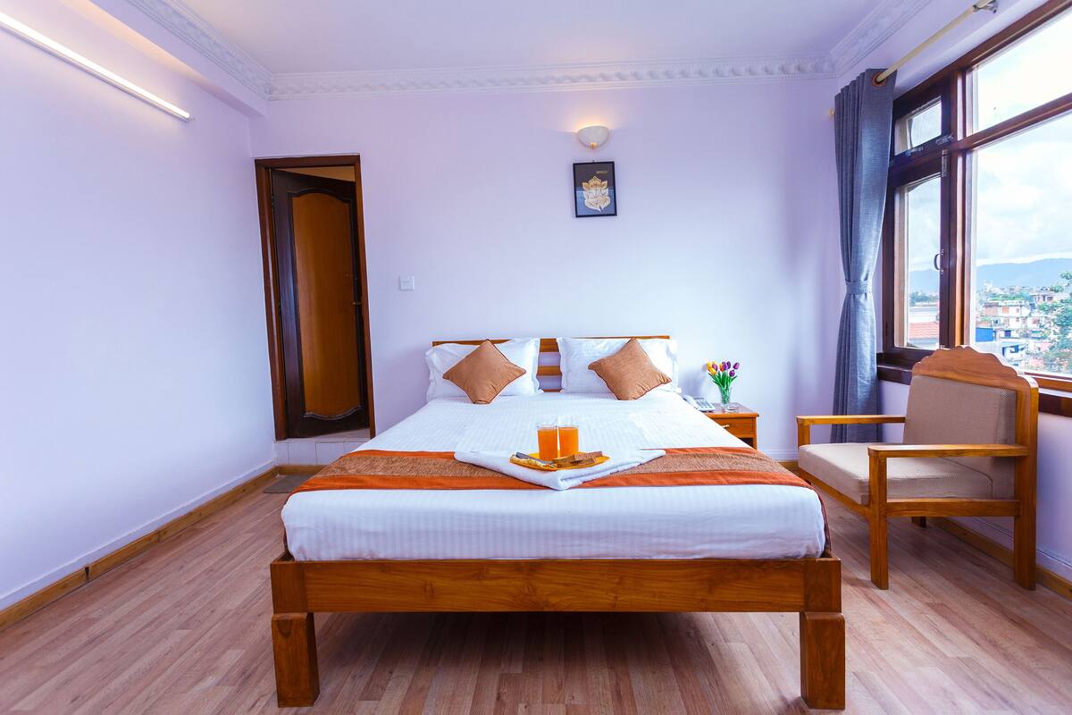 Una cama bien hecha, una bandeja con jugo de naranja, una silla de madera y vistas de la ciudad a través de una ventana.