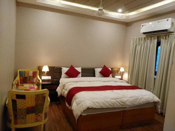 A hotel room with a bed, two bedside lamps, a chair with a quilt, an air conditioning unit, and curtains.