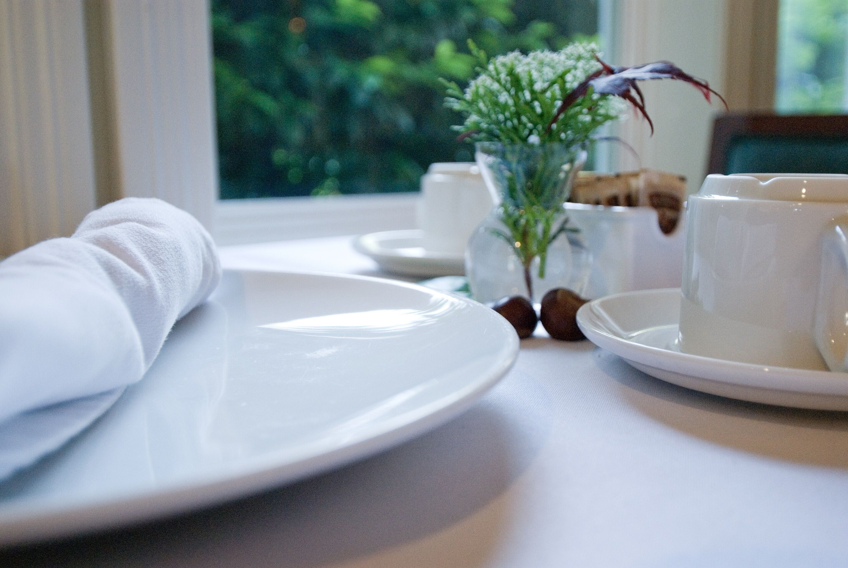 A neatly set dining table with white plates, cups, and a vase with flowers.