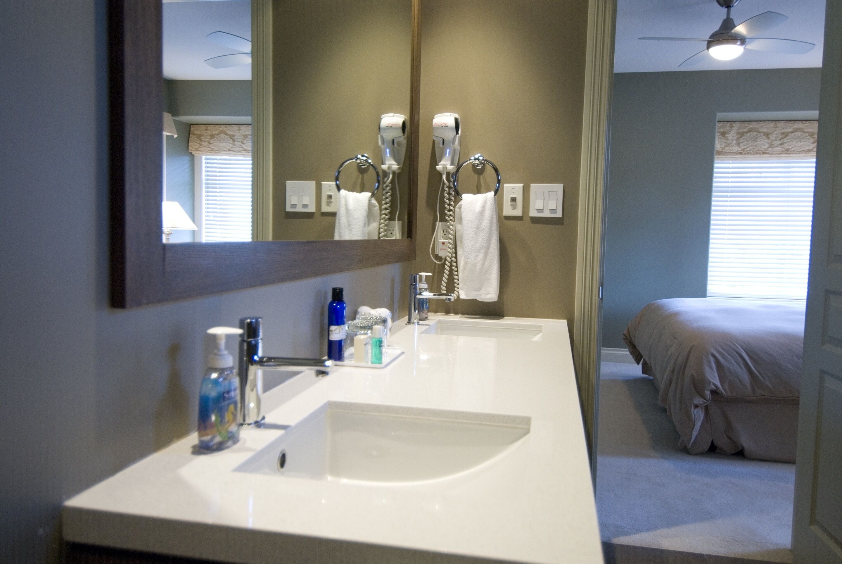 A bathroom with a large mirror, two sinks, a ceiling fan, and a bed in the adjacent room.