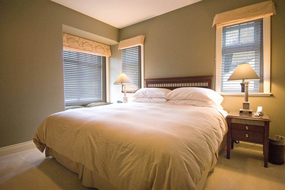 Een slaapkamer met een groot bed, twee bedlampjes, een nachtkastje en een raam met jaloezieën.