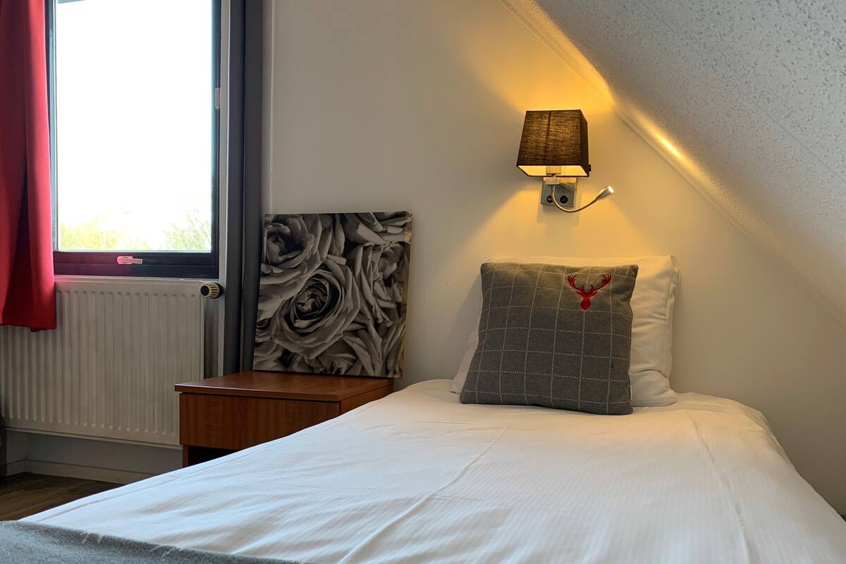 Una cama bien hecha con sábanas blancas, un almohadón gris con un emblema rojo y una lámpara fijada a la pared.