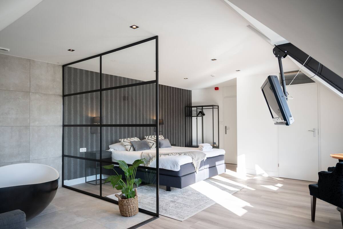 Un dormitorio moderno con una cama grande, un separador de vidrio, una silla negra y una planta.