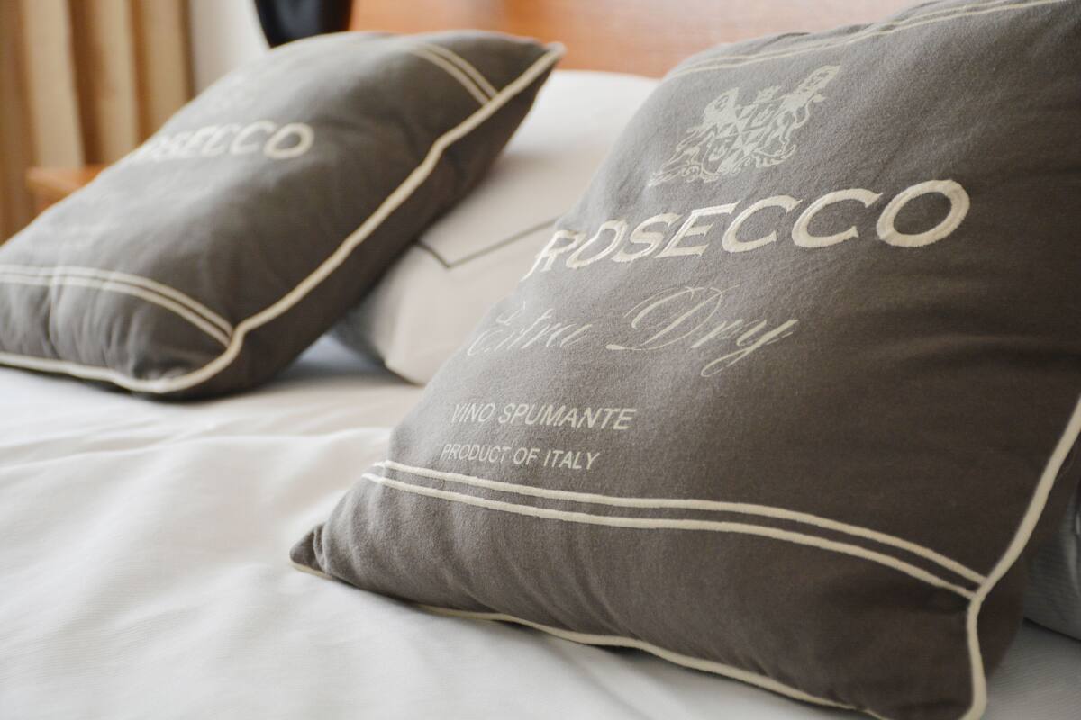 Dos almohadas con el texto "ROSECCO" y "VINO SPUMANTE PRODUCTO DE ITALIA" sobre una cama.