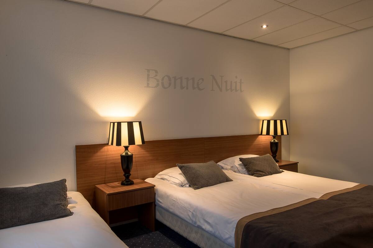 Habitación de hotel con dos camas, mesitas de noche y lámparas, y una pared con el texto "Bonne Nuit".