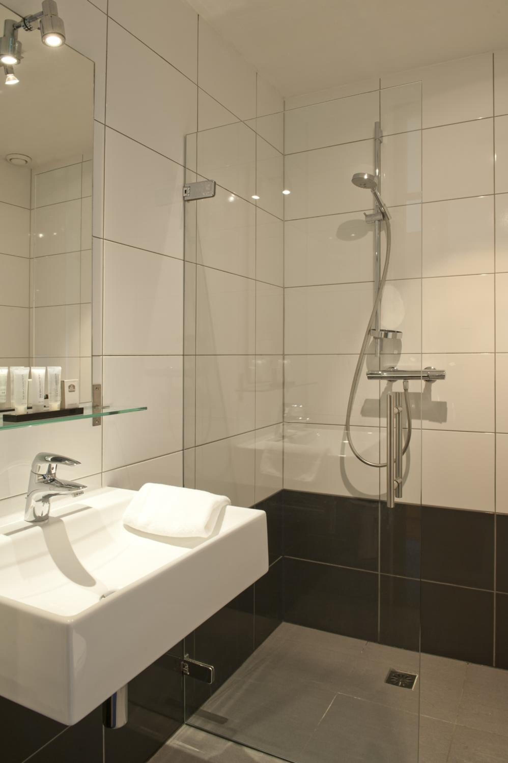 Une salle de bain moderne avec un lavabo blanc, une étagère en verre et une douche équipée d’un pommeau de douche à main.