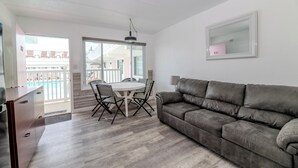 A modern living room with a grey sofa, a dining table with chairs, and a large window. - Alante Oceanfront Motel (North Wildwood)