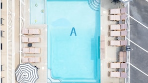 An aerial view of a pool area with a large 'A' sign, multiple lounge chairs, umbrellas, and a seating area with a sofa. - Alante Oceanfront Motel (North Wildwood)