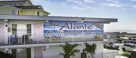 Un hôtel en bord de mer appelé «
Alante
», orné d’une fresque représentant des chaises de plage et un parasol, avec une clôture blanche et plusieurs voitures garées.