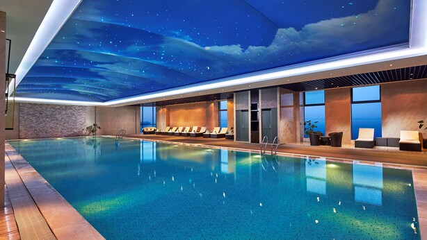 Una piscina interior moderna con techo que simula un cielo estrellado, sillones de descanso y vista al océano.