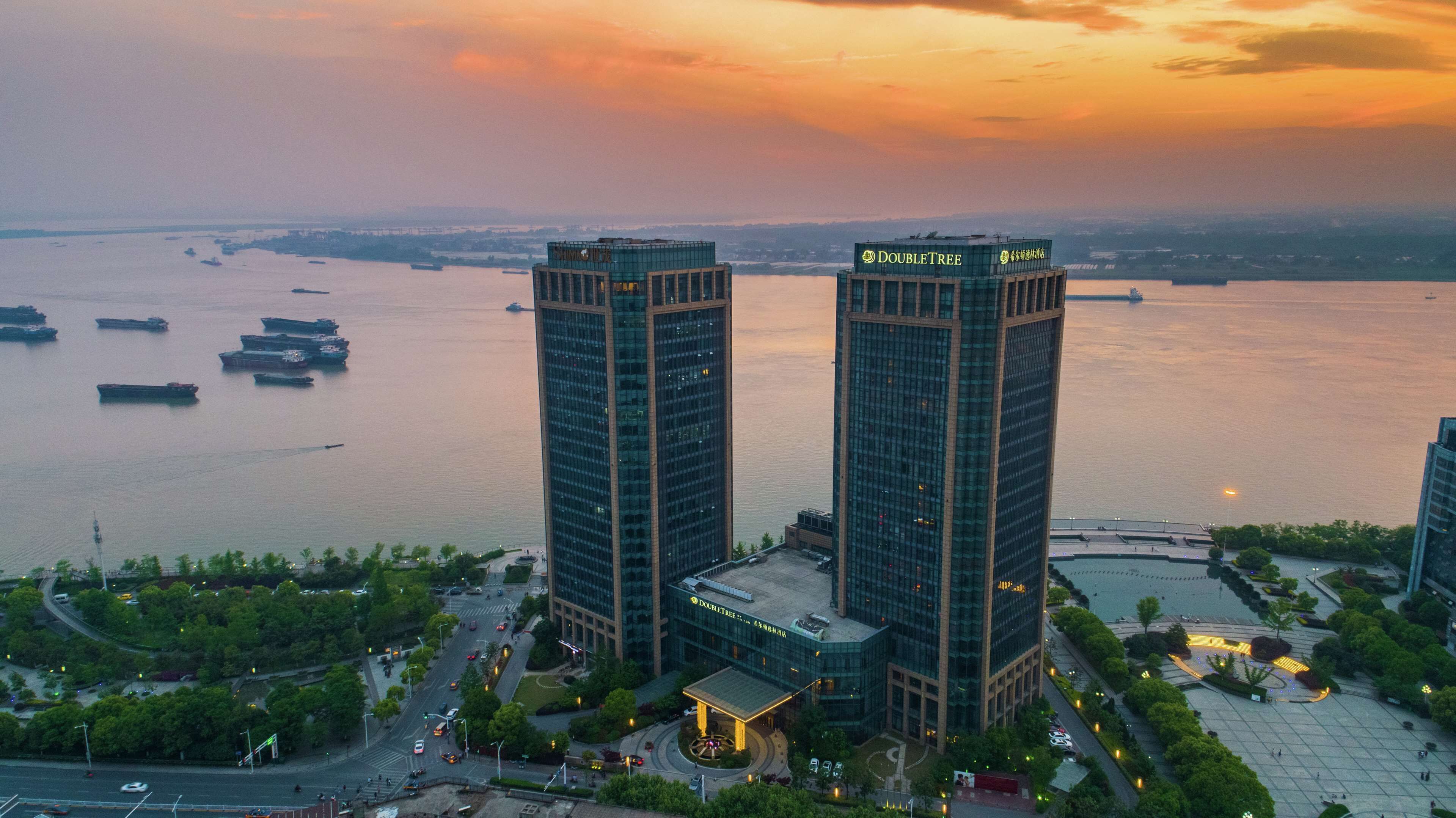 Un immeuble de grande hauteur, reconnaissable à son grand panneau « DOUBLETREE », entouré d’un espace paysager agrémenté d’arbres et offrant une vue sur le bord de l’eau.