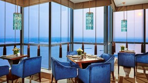 Un restaurante con amplios ventanales que ofrecen vistas al océano, sillas tapizadas en azul y candelabros de cristal colgantes.