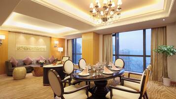 A hotel room with a dining table set for six, a sofa with cushions, a large window with a city view, and a chandelier.