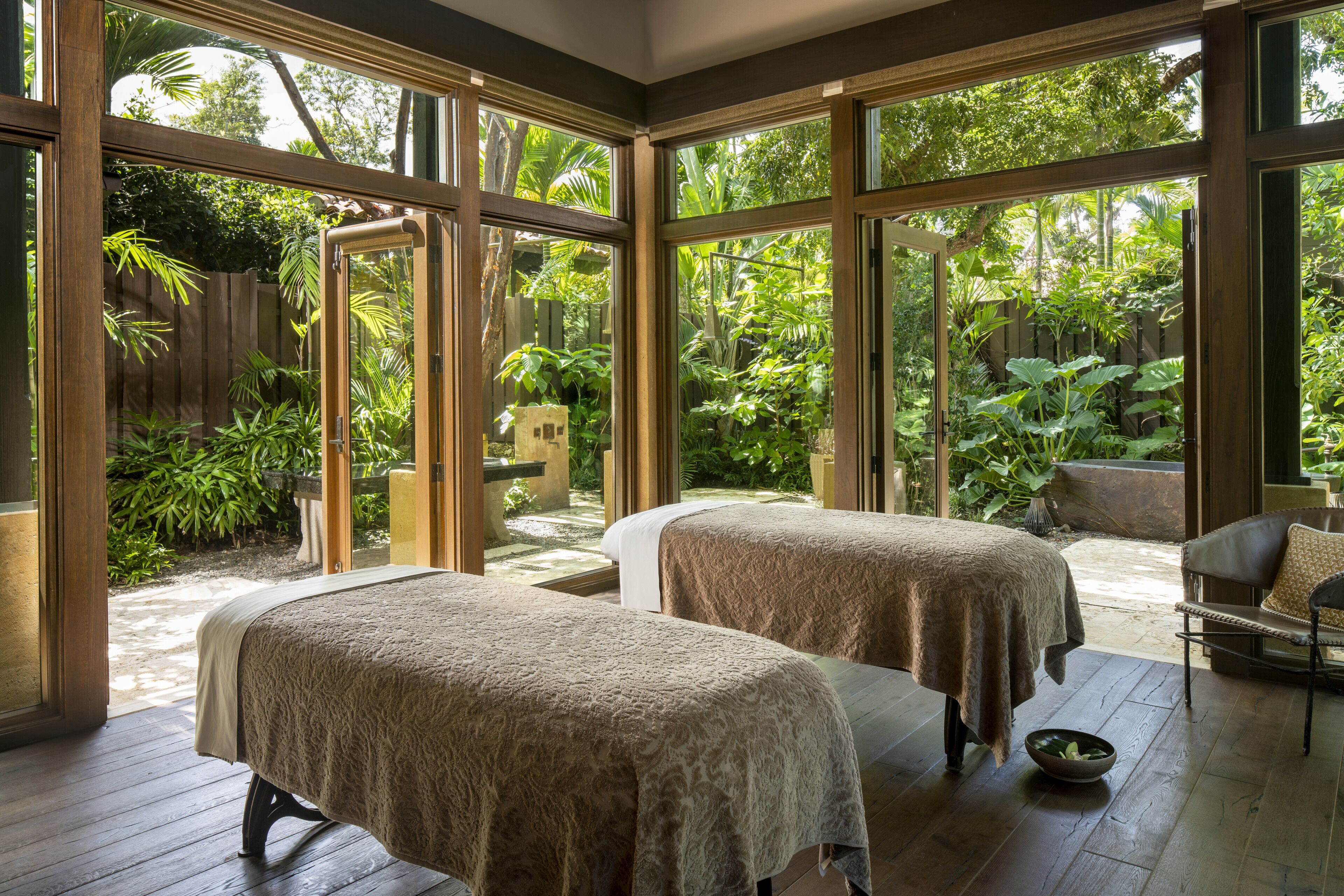 A room with two massage tables, large windows, and a view of a lush garden.