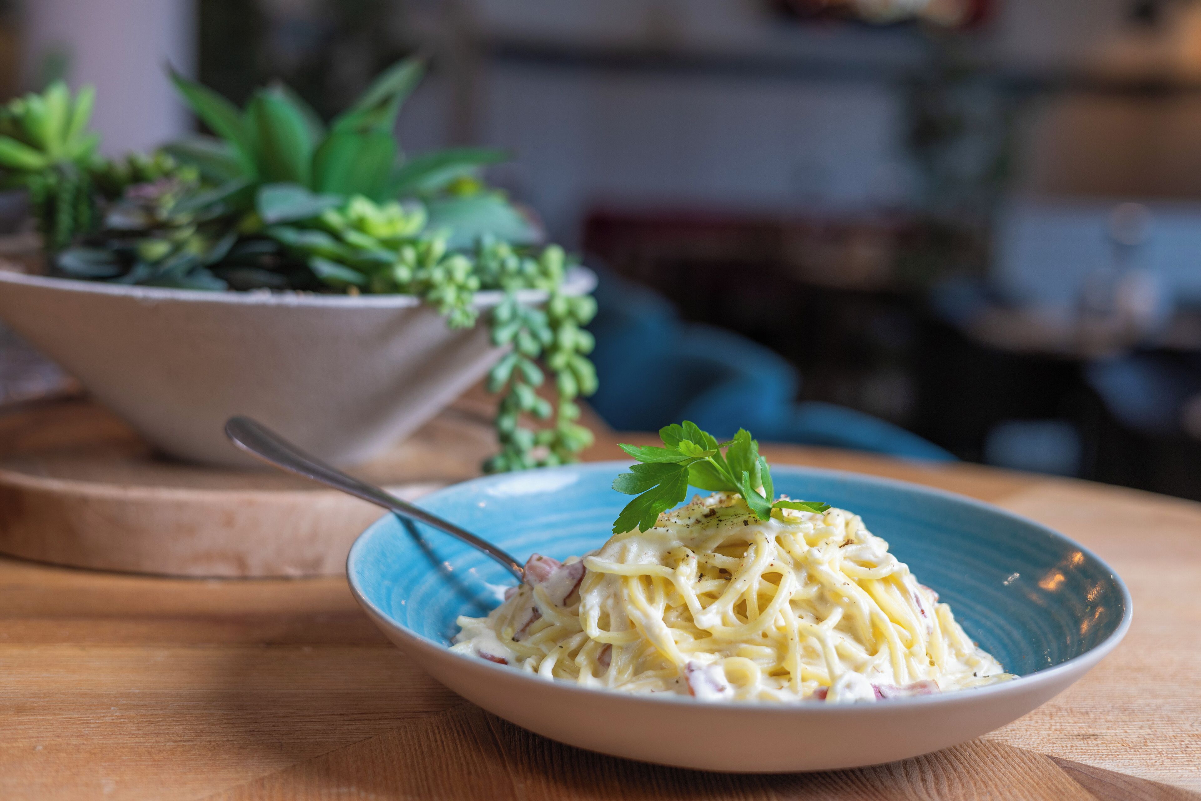 A bowl of pasta with a fork on a wooden table, with a potted plant in the background.