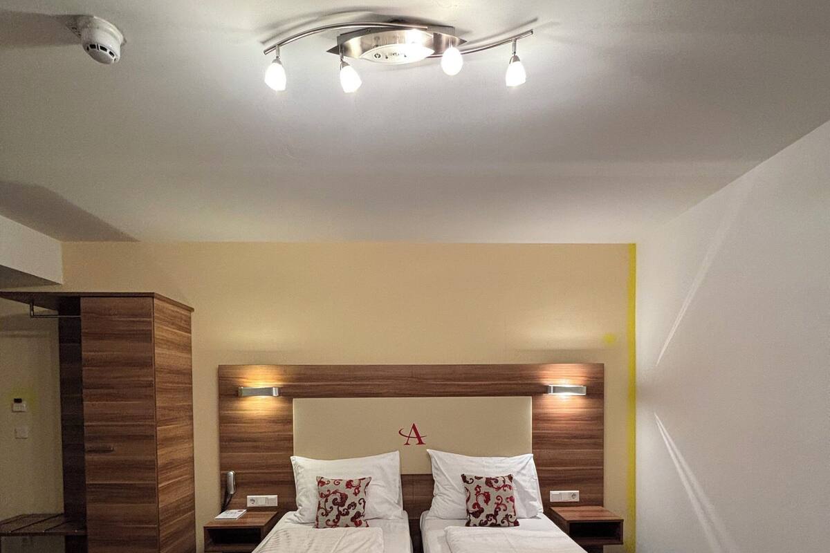 A hotel room with two beds, a wooden headboard, a ceiling light fixture, and a red towel on each bed.