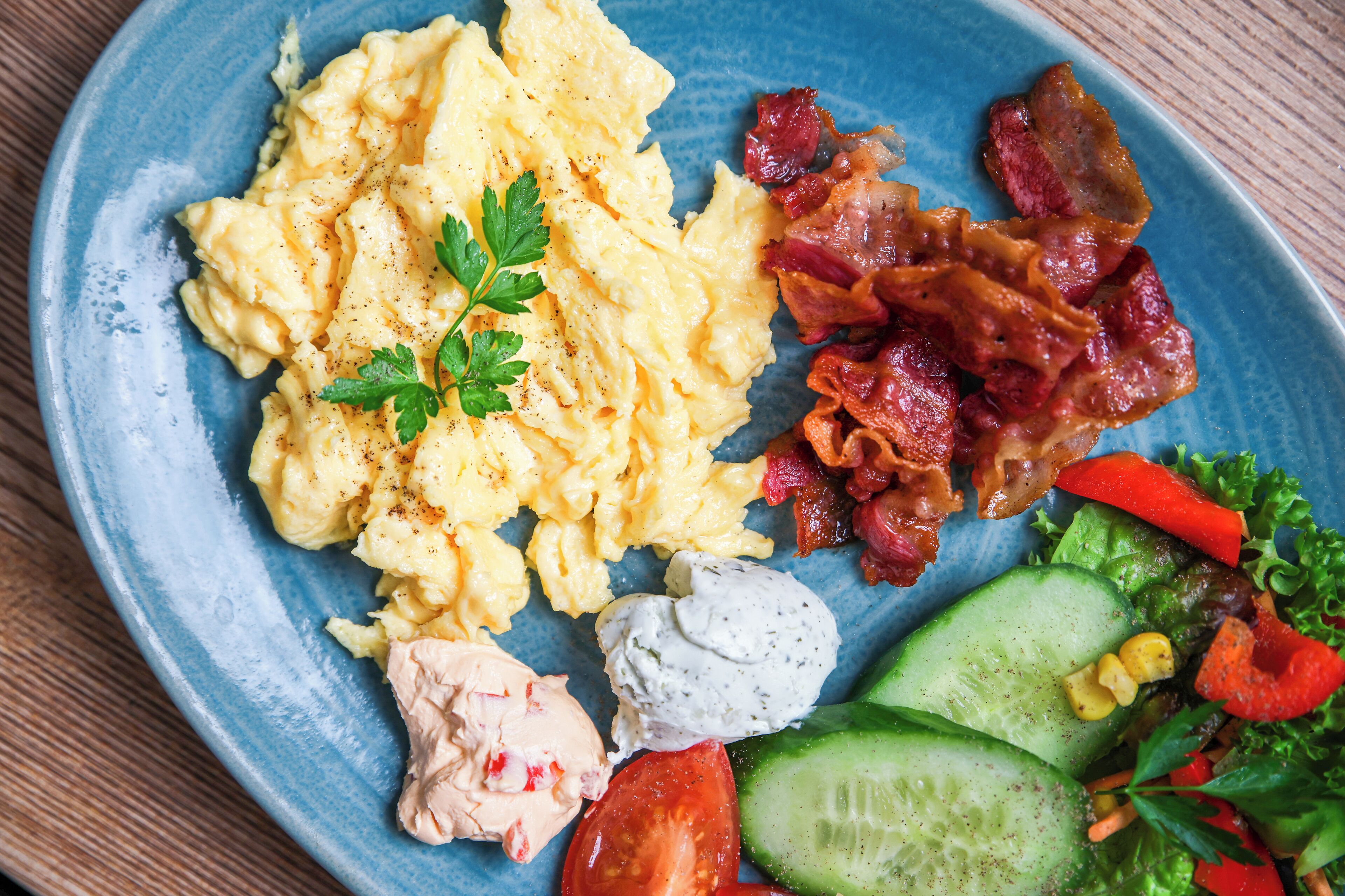 Scrambled eggs, bacon, a salad with cucumber, tomato, and red peppers, and a creamy sauce on a blue plate.