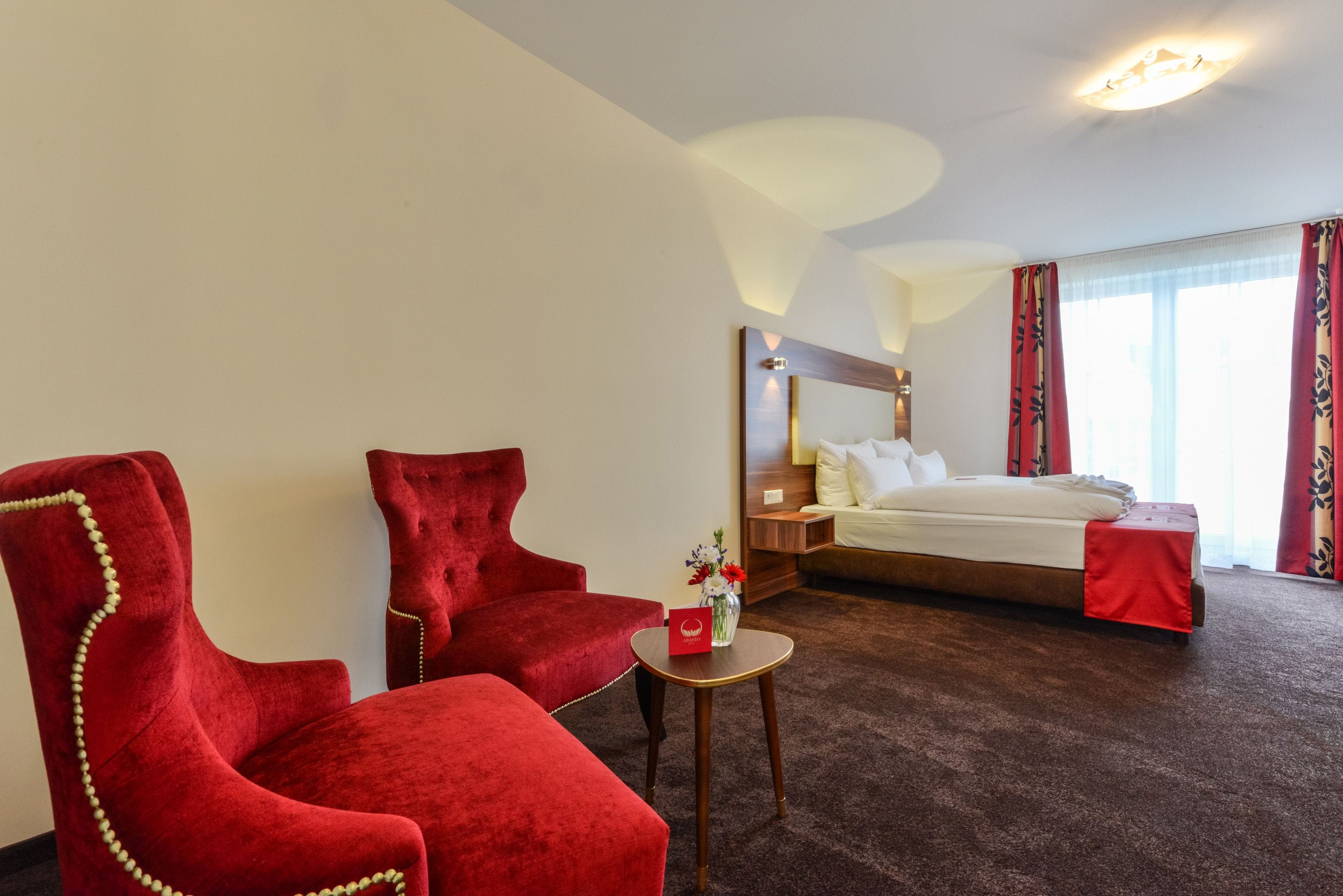 Een hotelkamer met een groot bed, twee rode fauteuils, een klein rond tafeltje en een nachtkastje met een lamp.