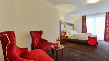 Een hotelkamer met een groot bed, twee rode fauteuils, een klein rond tafeltje en een nachtkastje met een lamp.