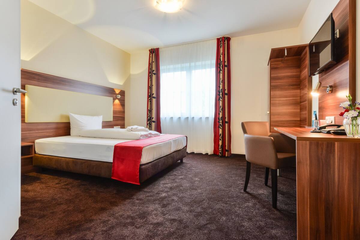A hotel room with a bed, a desk, a chair, a TV, and a window with curtains.