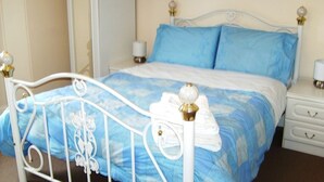 A bed with a white metal frame and blue bedding, a bedside table with a lamp, and a nightstand with drawers. - Hamsteels Hall (Durham)