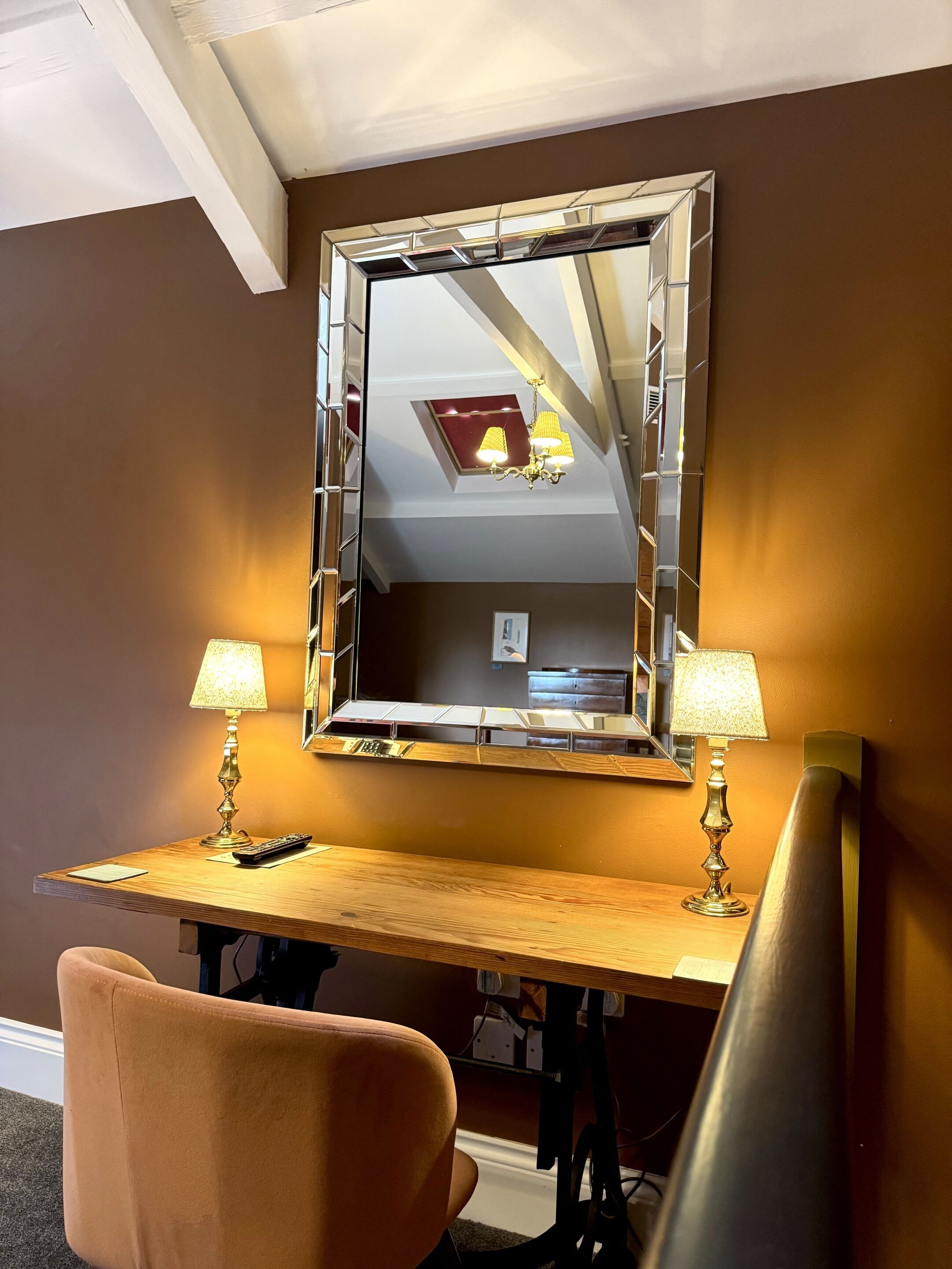 A hotel room with a wooden desk, two lamps, a mirror, and a chair.