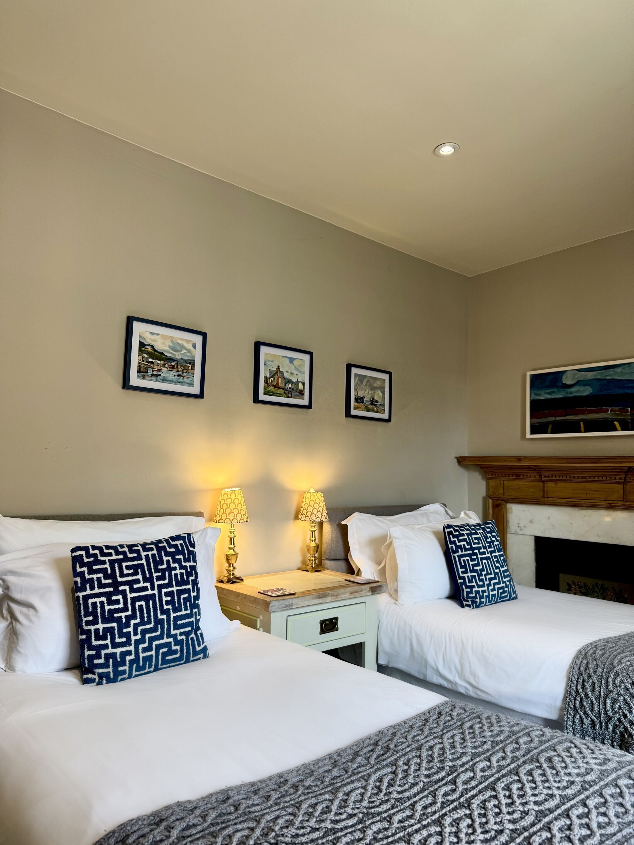 A hotel room with two beds, a nightstand with lamps, a fireplace, and three framed pictures on the wall.