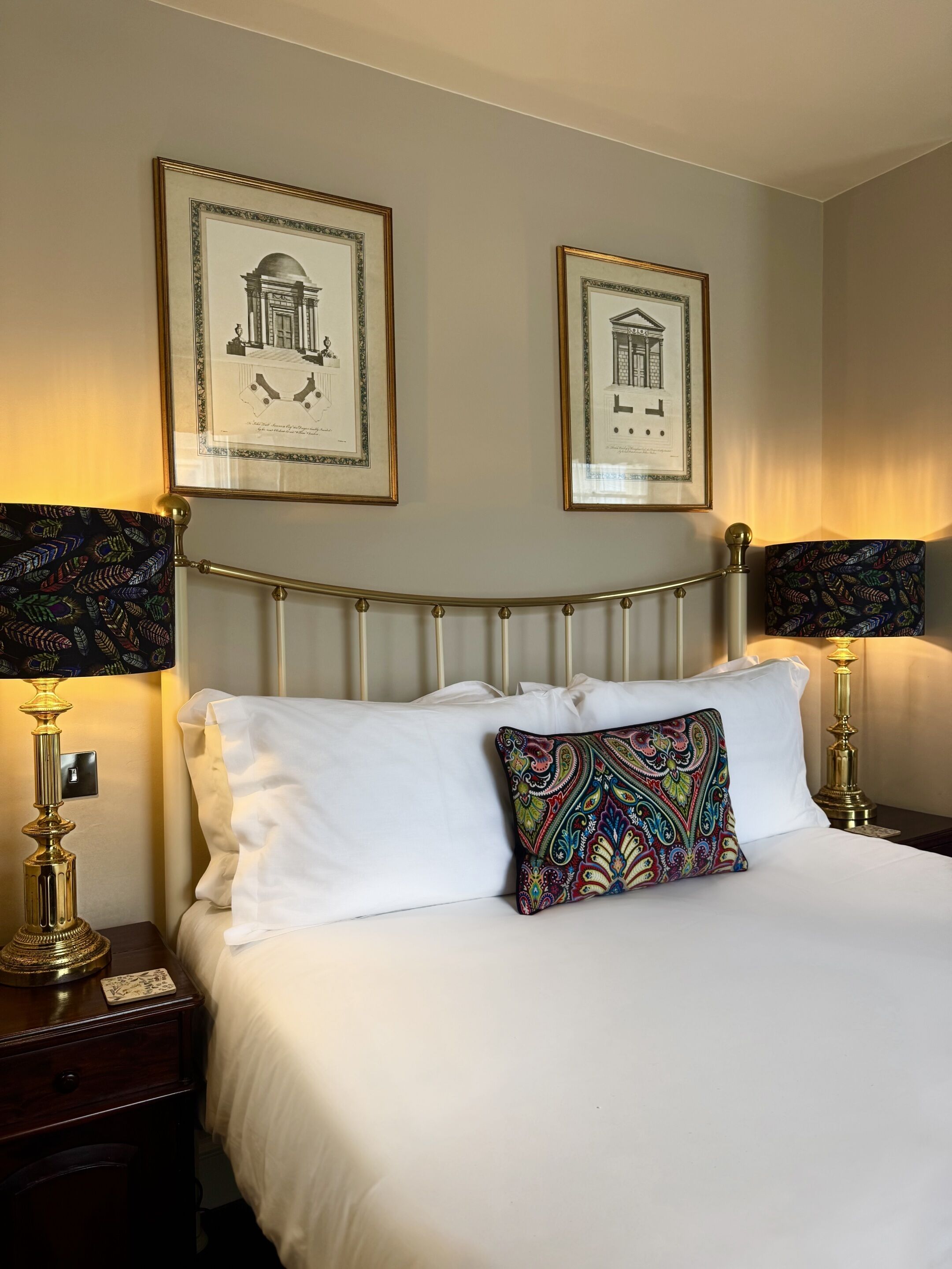 A neatly made bed with white linens and a colorful paisley pillow, flanked by two gold-framed lamps with patterned shades. Above the bed, there are two framed architectural drawings on the wall.