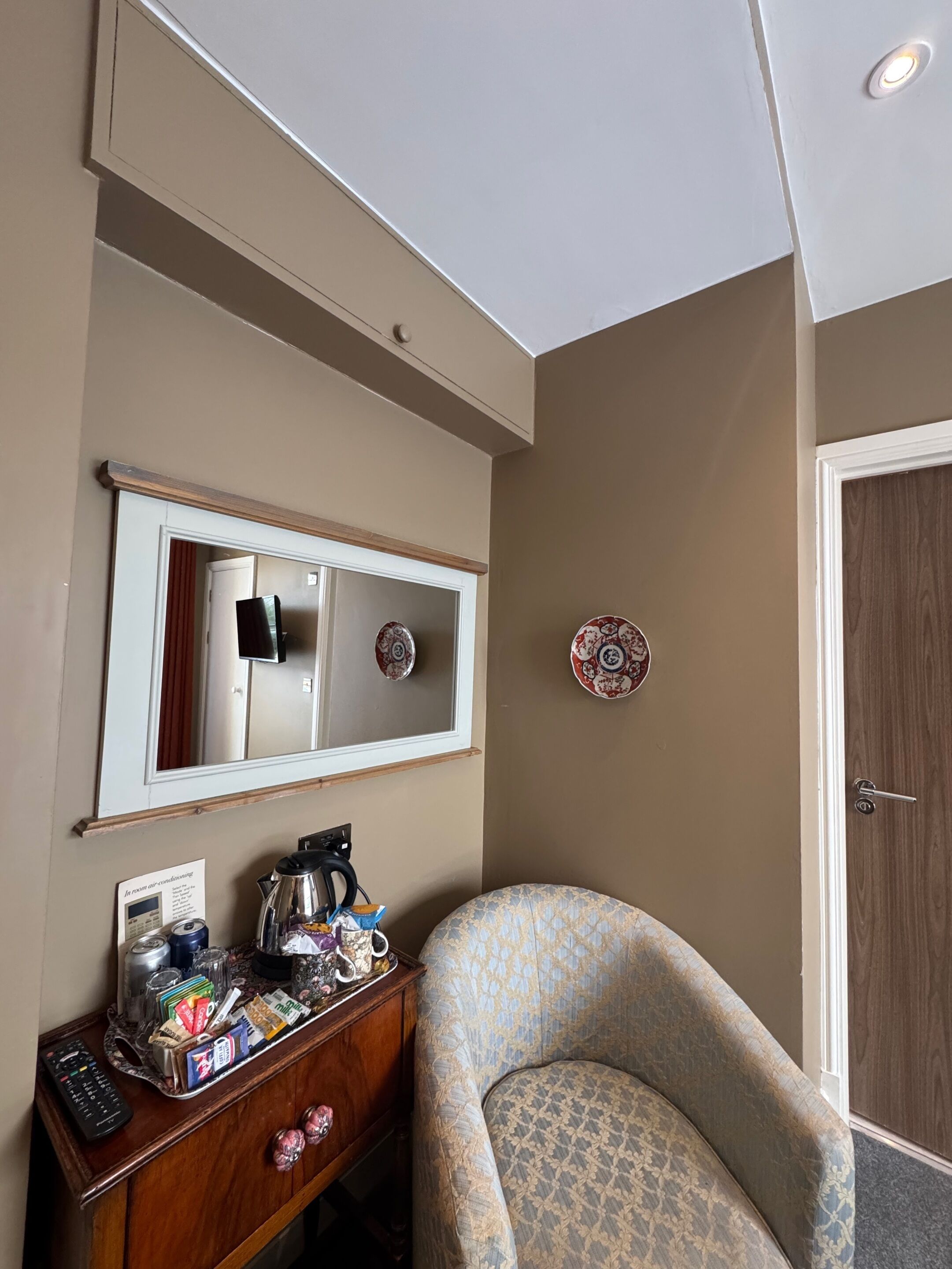 Una habitación de hotel con un aparador de madera, un sillón estampado, un espejo y una puerta.