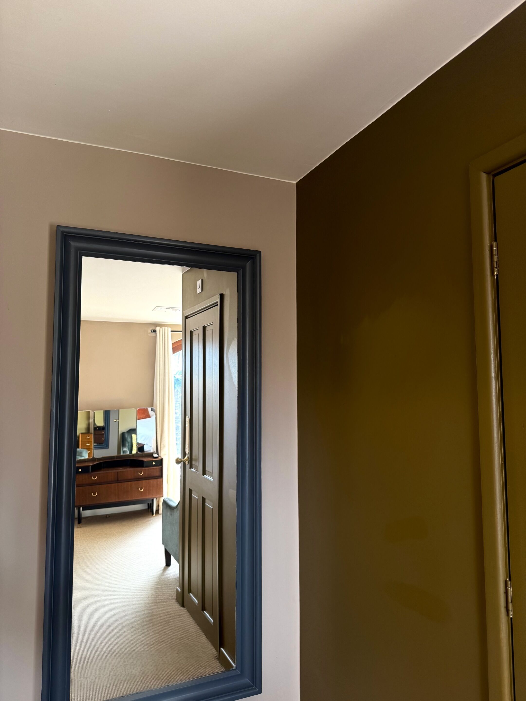 A hallway with a mirror reflecting a room with a dresser and a door.