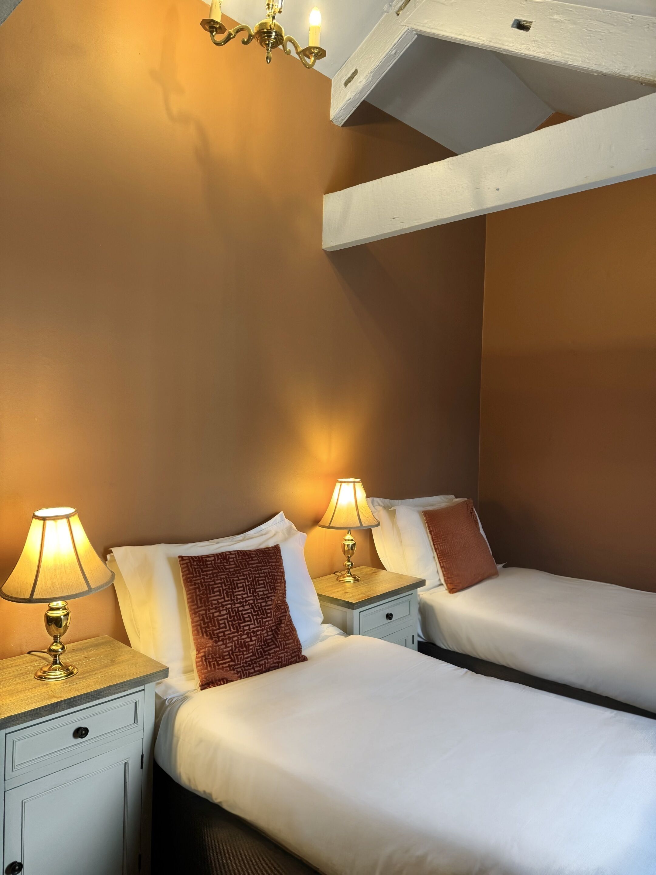 A hotel room with two single beds, each with a bedside table and lamp. The room has a wooden ceiling and a warm wall color.