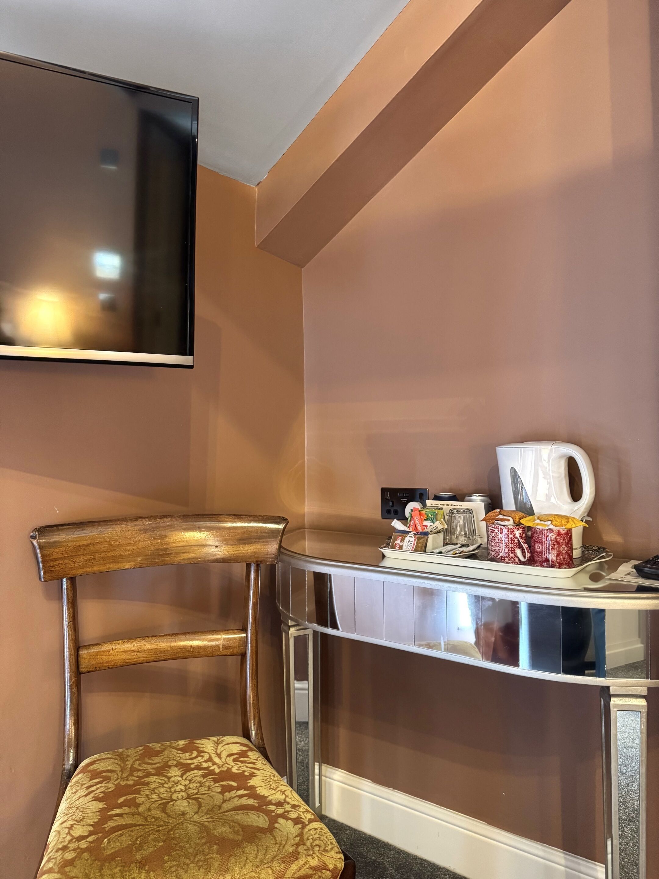 A hotel room with a wooden chair, a small table with a coffee maker, and a TV mounted on the wall.