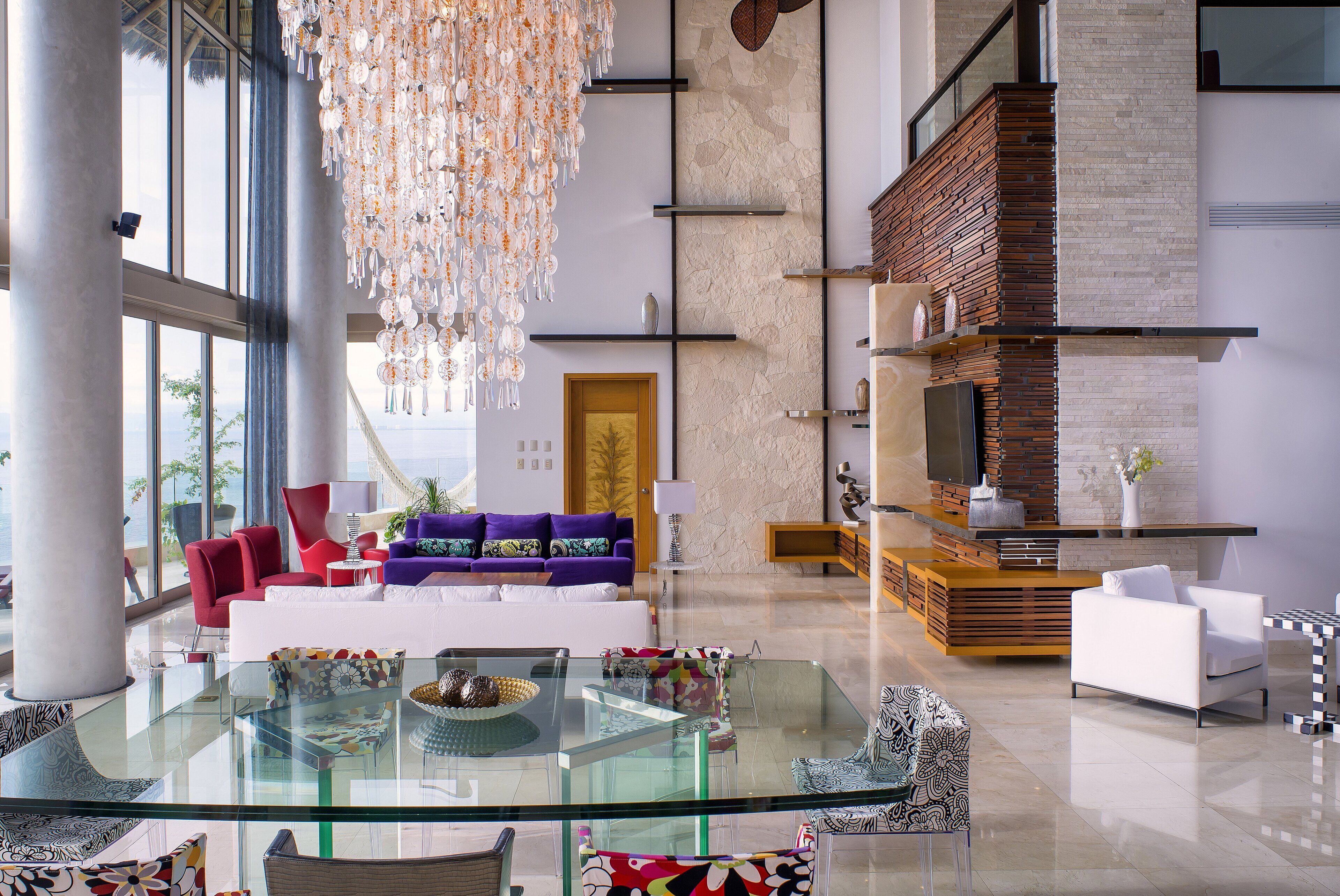 Un amplio salón con una mesa de centro de vidrio, sofás rojos y morados, una chimenea y ventanas grandes.