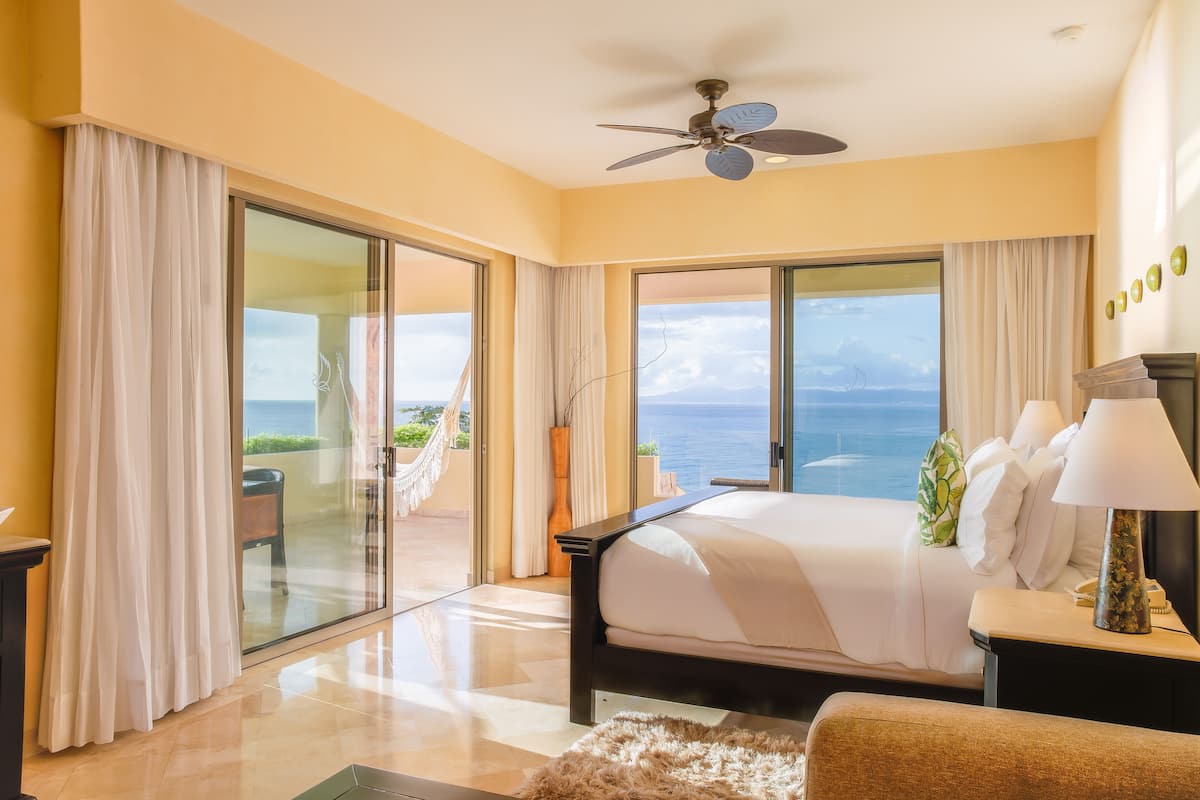 A bedroom with a large bed, a ceiling fan, and a view of the ocean through large windows.