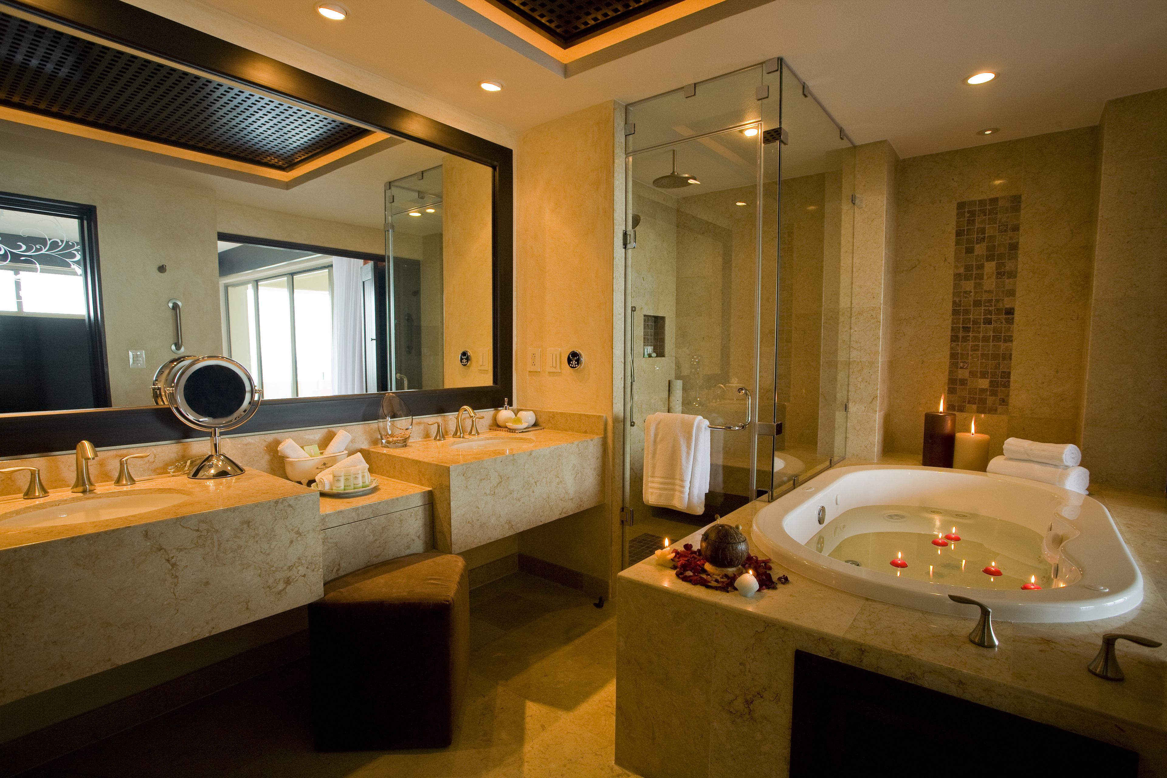 A modern bathroom with dual sinks, a large mirror, a glass-enclosed shower, and a bathtub with candles.