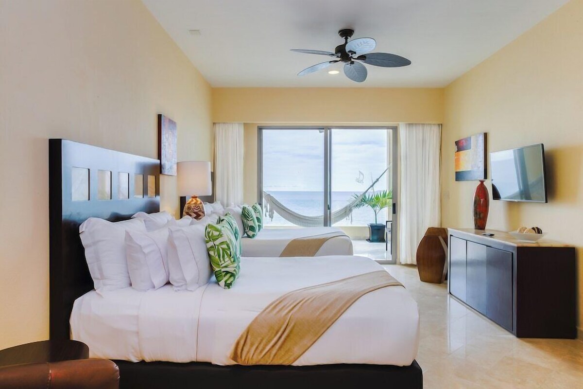 A hotel room with a large bed, a ceiling fan, a TV, a desk, and a view of the ocean.