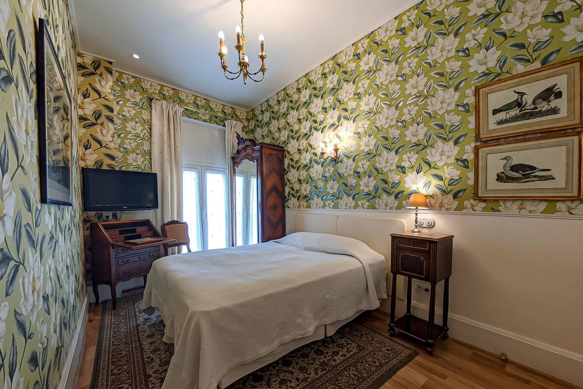 Ein Schlafzimmer mit einem Bett, einem Schreibtisch, einem Fernseher, einer Kristallleuchter und blumigem Wandpapier.