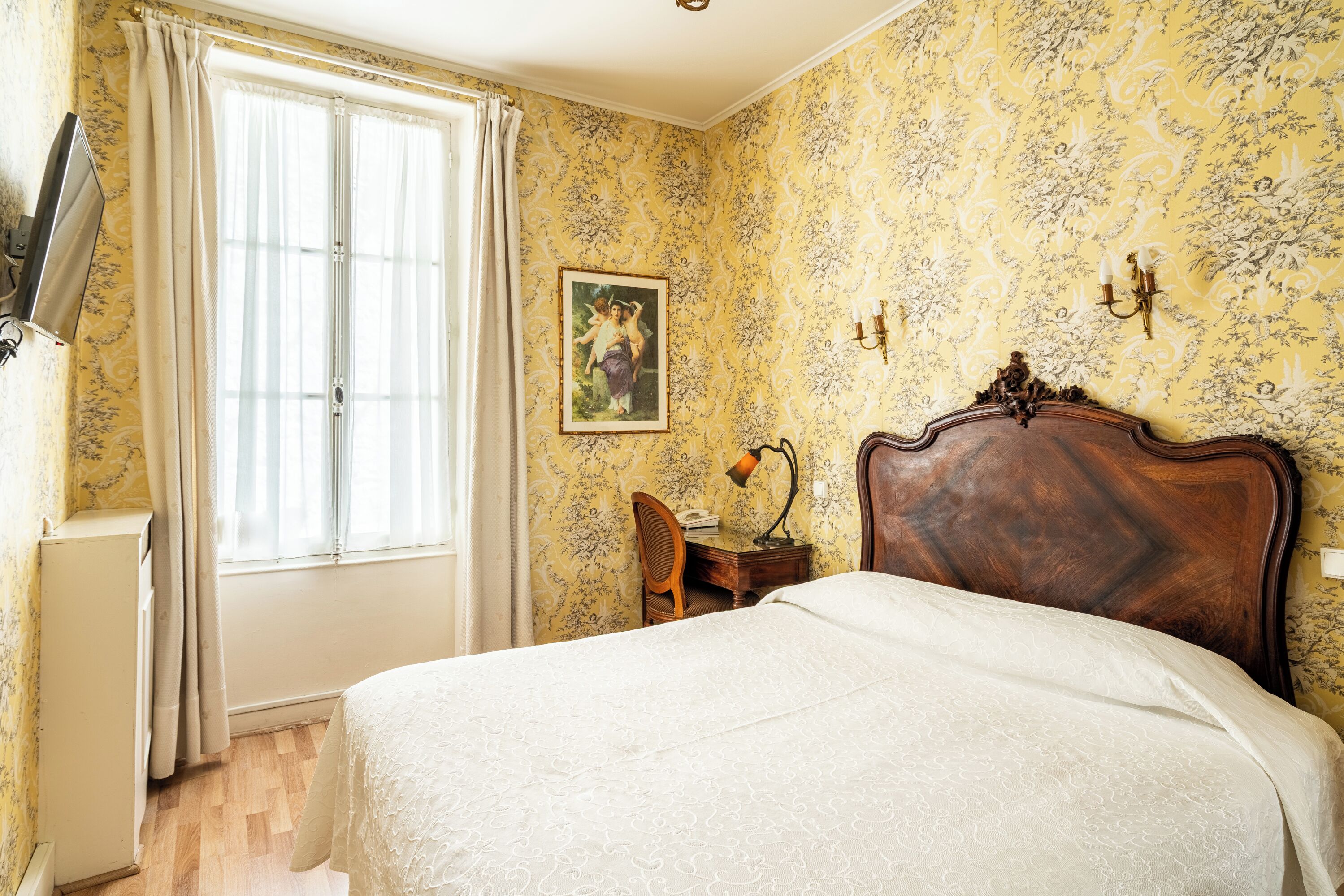 A bedroom with a wooden bed, a desk with a lamp, a chair, a painting on the wall, and a window with curtains.