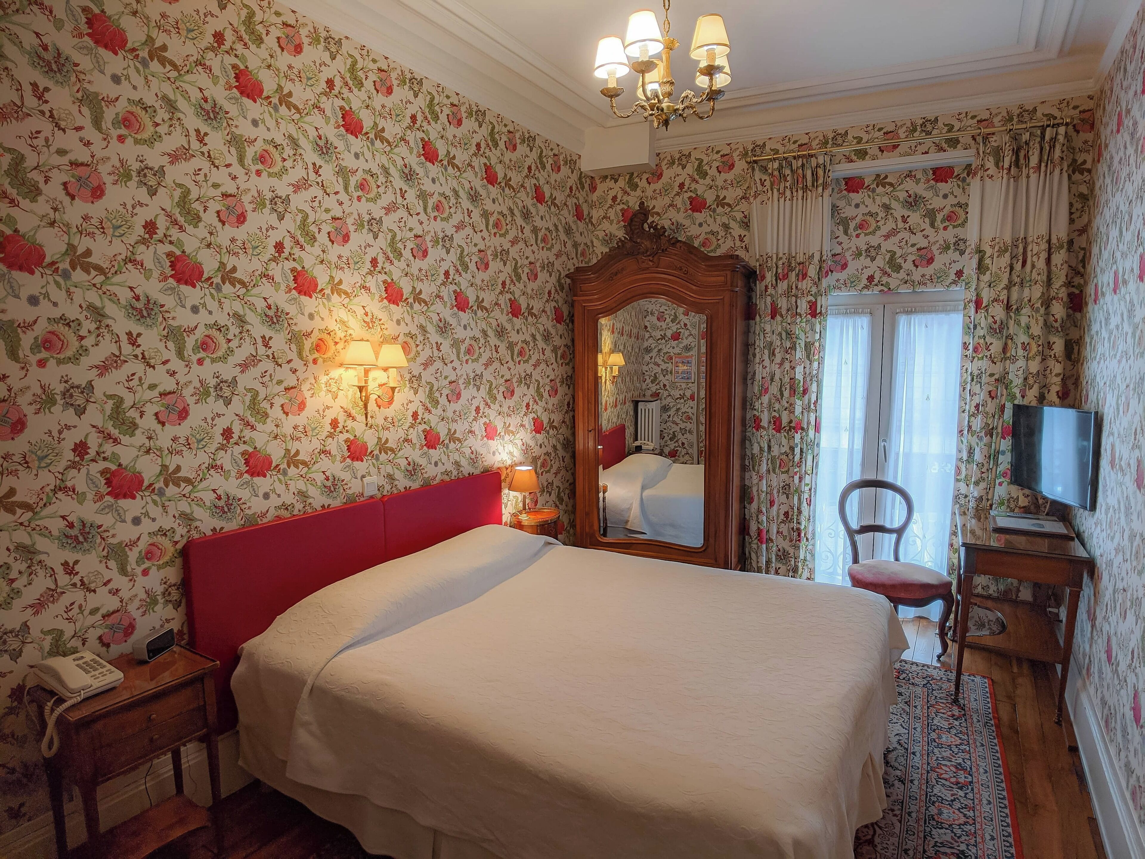 Un dormitorio con papel tapiz floral, un cabecero rojo, un espejo grande, una mesita de noche con lámpara, una silla y un televisor.