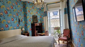 Een slaapkamer met bloemenbehang, een kroonluchter, een bed, een houten dressoir, een stoel en uitzicht op een gebouw door een balkondeur.