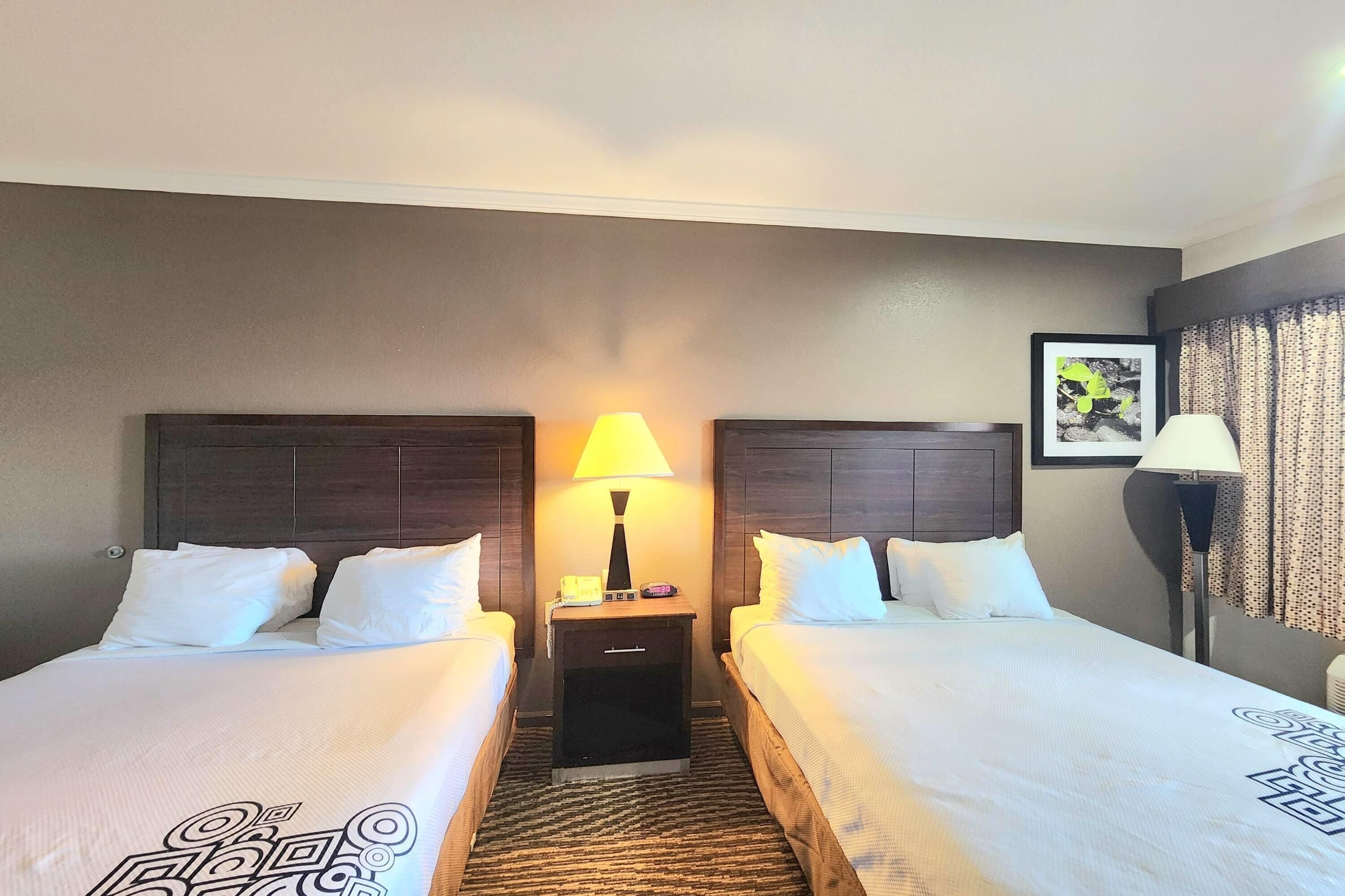 A hotel room with two beds, a nightstand with a lamp, a clock, and a framed picture on the wall.