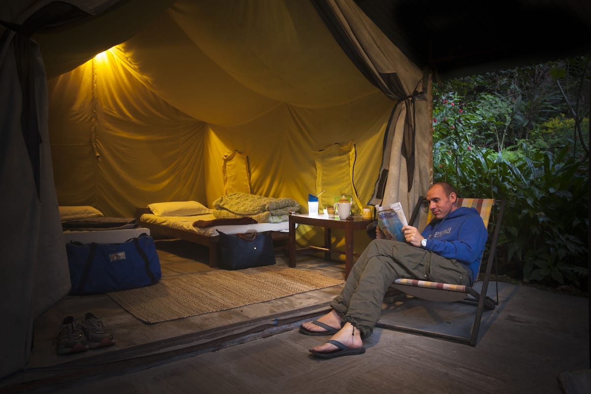 Tenda com uma cama, mesa com uma xícara e um livro, e uma pessoa sentada em uma cadeira lendo.