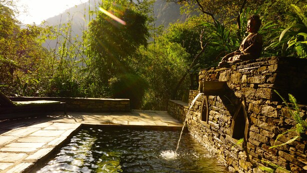 A stone water feature with a statue, surrounded by lush greenery and a stone pathway. - The Last Resort (Listikot)